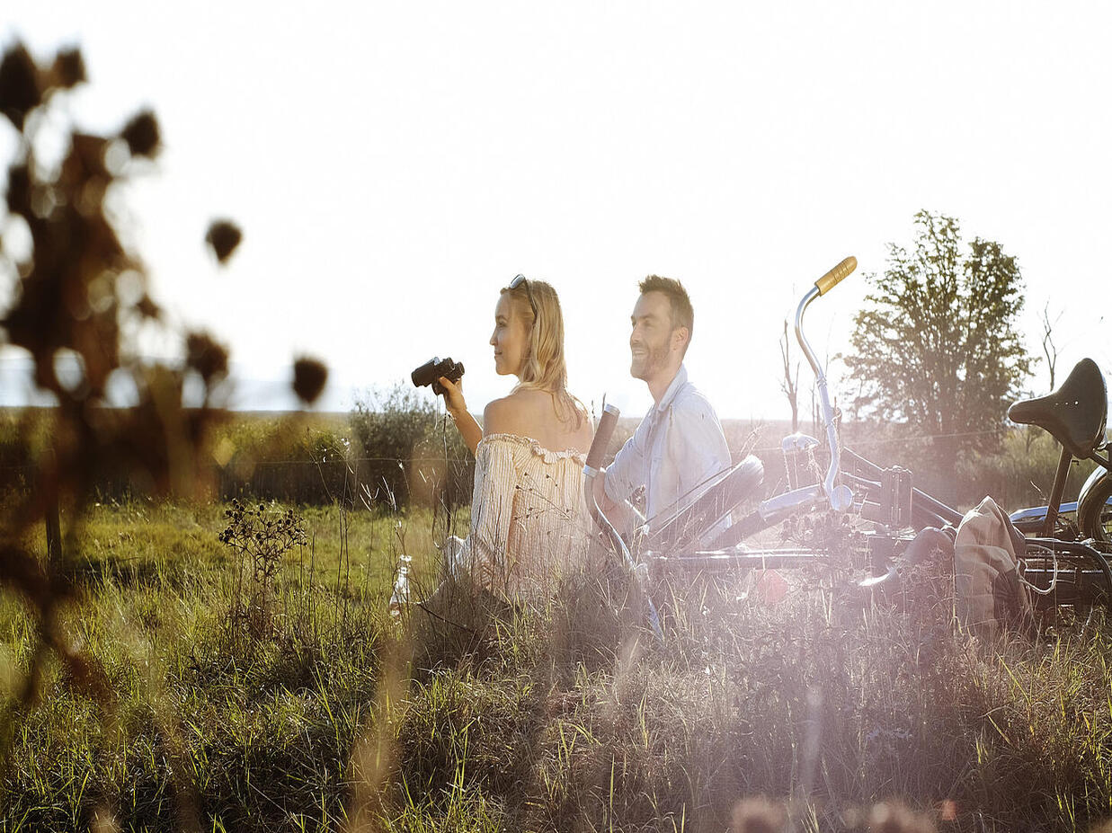 Zwei Erwachsene sitzen mit Fahrrädern im hohen Gras einer weiten Landschaft, die tief stehende Sonne scheint von hinten; eine Person hält ein Fernglas und blickt in die Natur.