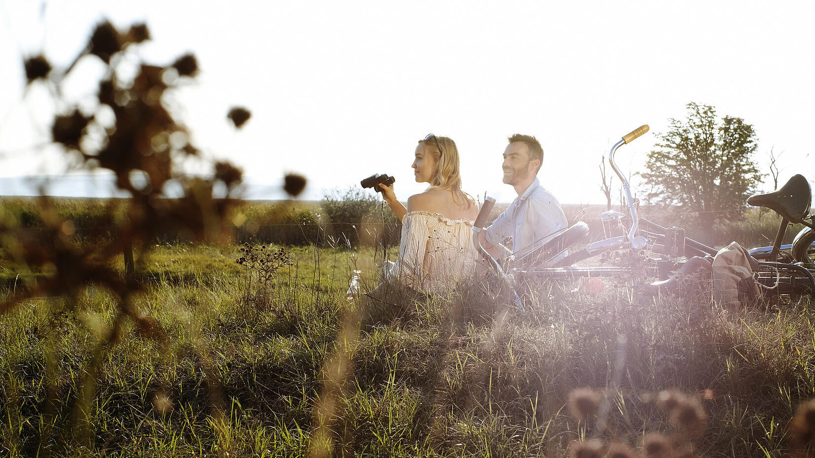 Zwei Erwachsene sitzen mit Fahrrädern im hohen Gras einer weiten Landschaft, die tief stehende Sonne scheint von hinten; eine Person hält ein Fernglas und blickt in die Natur.
