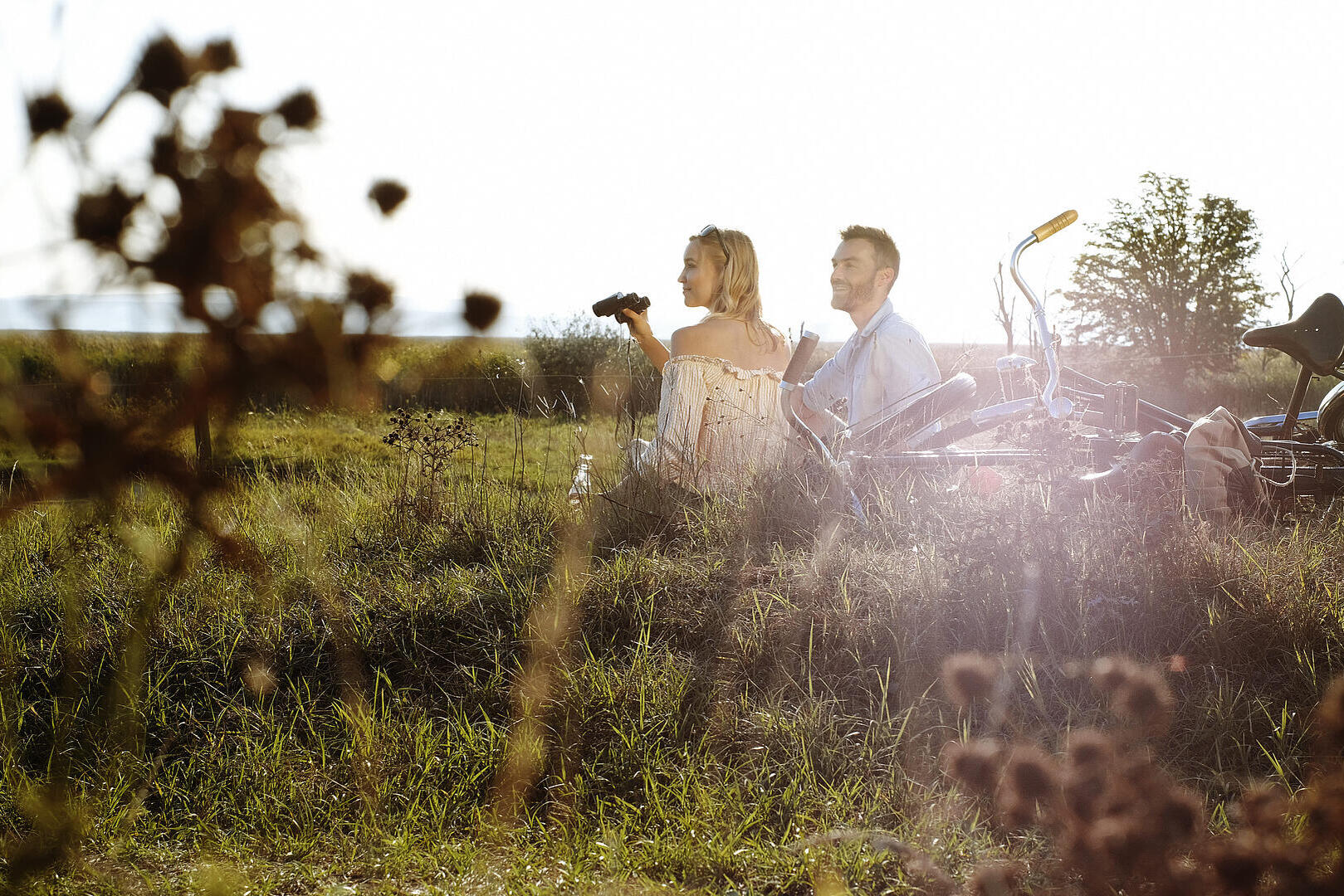 Zwei Erwachsene sitzen mit Fahrrädern im hohen Gras einer weiten Landschaft, die tief stehende Sonne scheint von hinten; eine Person hält ein Fernglas und blickt in die Natur.