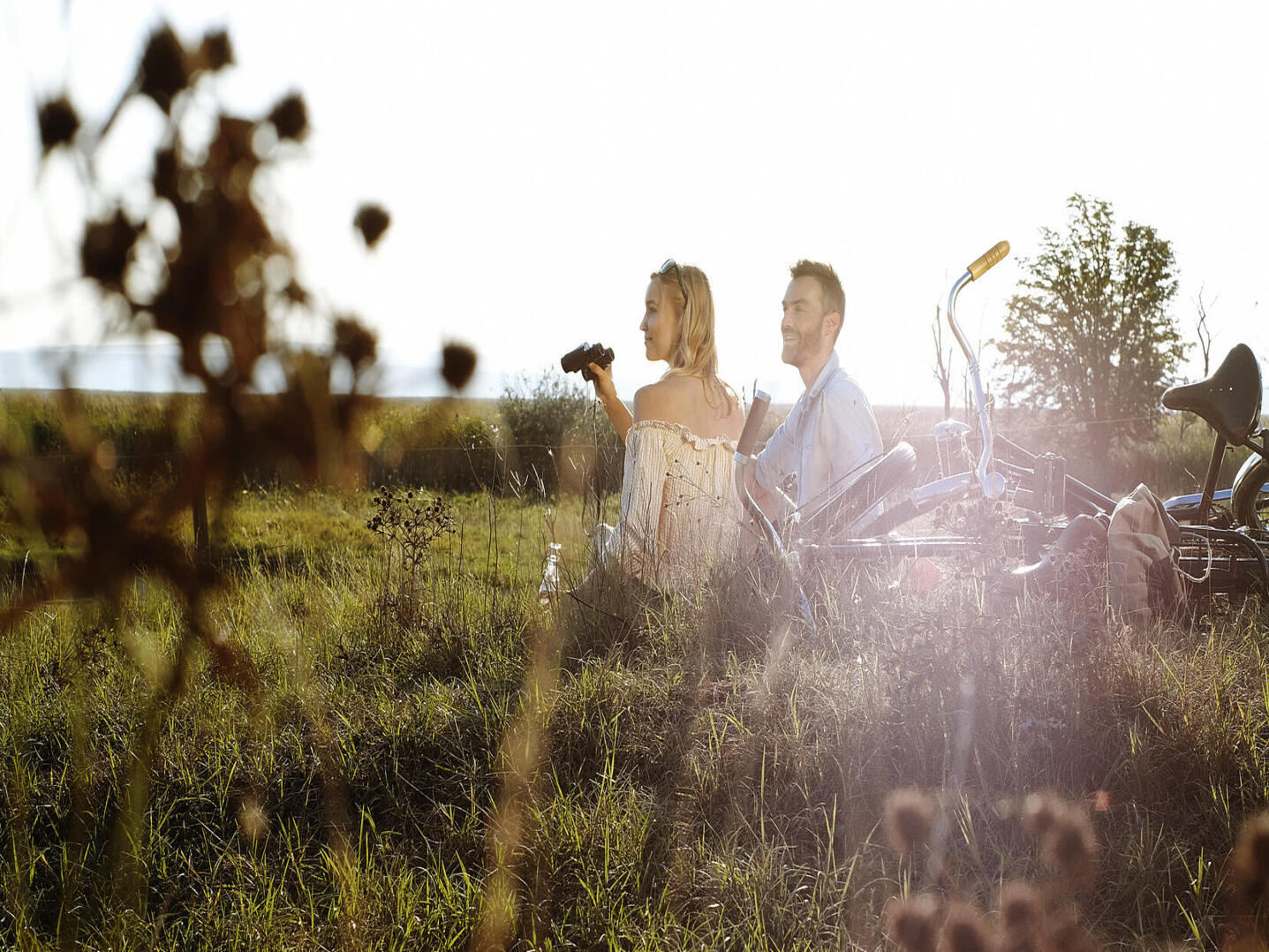 Zwei Erwachsene sitzen mit Fahrrädern im hohen Gras einer weiten Landschaft, die tief stehende Sonne scheint von hinten; eine Person hält ein Fernglas und blickt in die Natur.