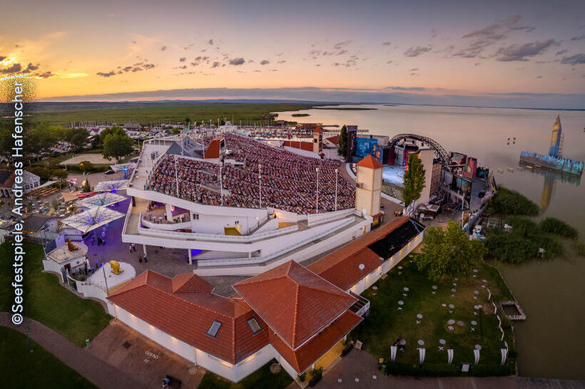 Luftaufnahme der Seefestspiele Mörbisch am Neusiedler See bei Sonnenuntergang, mit voll besetzter Zuschauertribüne und Seebühne am Wasser.
