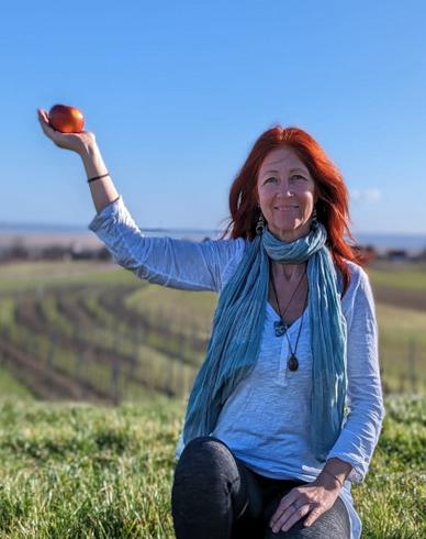 „Eine lächelnde Person mit roten Haaren kniet auf einer Wiese und hält einen roten Apfel in die Höhe. Im Hintergrund erstrecken sich Felder und Weinreben unter einem klaren blauen Himmel.“